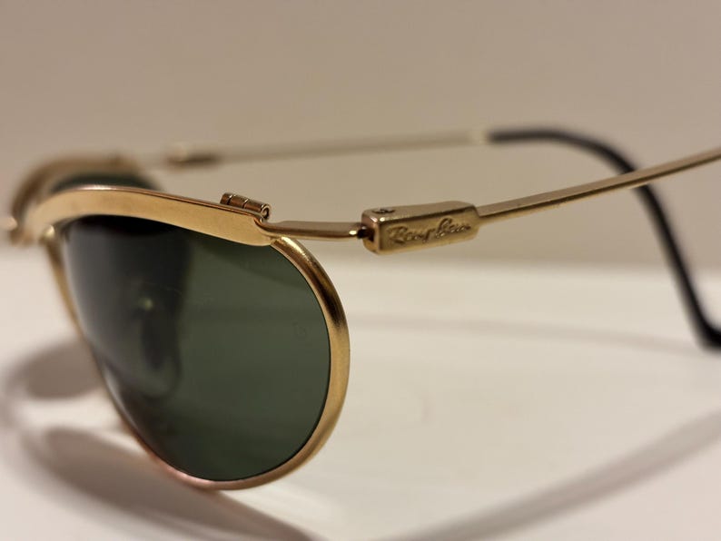 Puede incluir: Primer plano de gafas de sol con montura dorada y lentes verdes. El logotipo de Ray-Ban es visible en la patilla. Las gafas de sol tienen un dise&ntilde;o cl&aacute;sico con montura dorada y patillas negras.