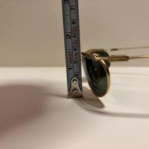 Puede incluir: Gafas de sol con montura dorada y lentes verde oscuro. La altura de las gafas, medida con una cinta m&eacute;trica met&aacute;lica, es de aproximadamente 5 cm. Las gafas tienen un estilo aviador cl&aacute;sico.