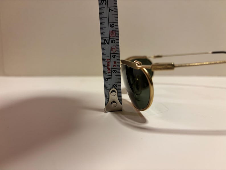 Puede incluir: Gafas de sol con montura dorada y lentes verde oscuro. La altura de las gafas, medida con una cinta m&eacute;trica met&aacute;lica, es de aproximadamente 5 cm. Las gafas tienen un estilo aviador cl&aacute;sico.