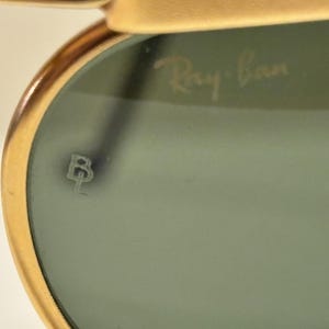 Puede incluir: Primer plano de gafas de sol Ray-Ban. La montura dorada rodea una lente te&ntilde;ida de verde. El logotipo de Ray-Ban es visible en la lente. Tambi&eacute;n hay un peque&ntilde;o emblema estilizado en la lente.