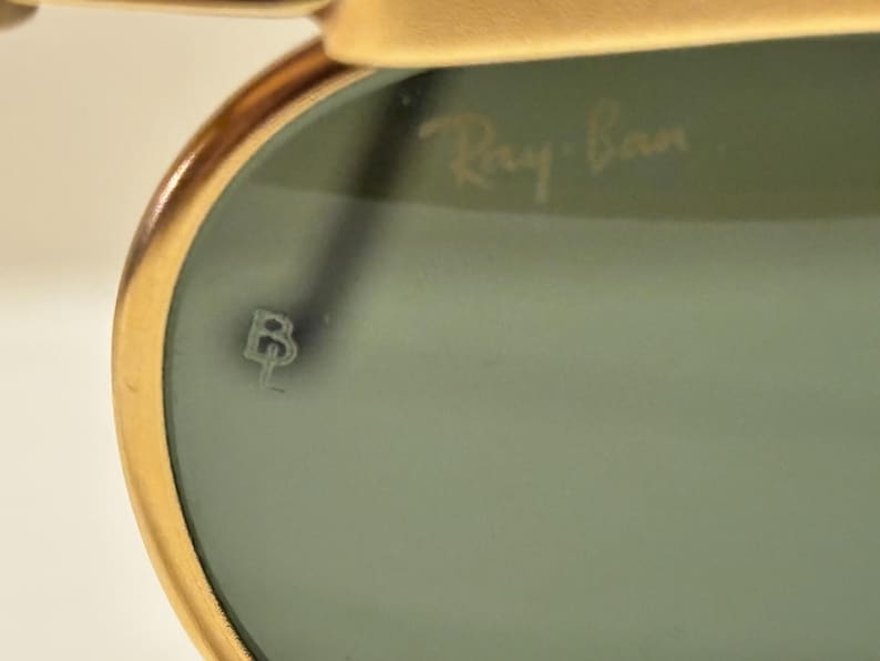 Puede incluir: Primer plano de gafas de sol Ray-Ban. La montura dorada rodea una lente te&ntilde;ida de verde. El logotipo de Ray-Ban es visible en la lente. Tambi&eacute;n hay un peque&ntilde;o emblema estilizado en la lente.