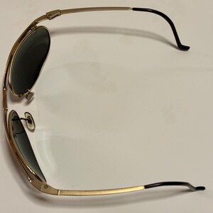 Puede incluir: Gafas de sol doradas con lentes oscuras. Las monturas tienen un estilo aviador cl&aacute;sico con doble puente y puntas de patillas negras. Las gafas de sol est&aacute;n en &aacute;ngulo, mostrando la forma y el dise&ntilde;o.