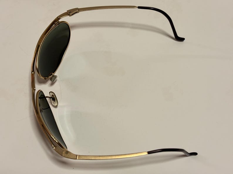 Puede incluir: Gafas de sol doradas con lentes oscuras. Las monturas tienen un estilo aviador cl&aacute;sico con doble puente y puntas de patillas negras. Las gafas de sol est&aacute;n en &aacute;ngulo, mostrando la forma y el dise&ntilde;o.