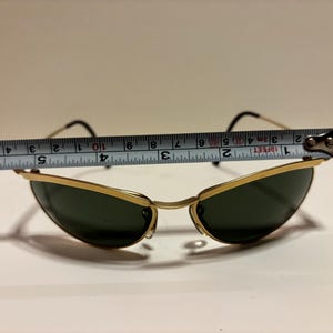 Puede incluir: Gafas de sol con montura dorada y lentes verde oscuro. Las gafas de sol se miden con una cinta m&eacute;trica, mostrando un ancho de aproximadamente 12,7 cm. Las patillas son negras y la montura es dorada.