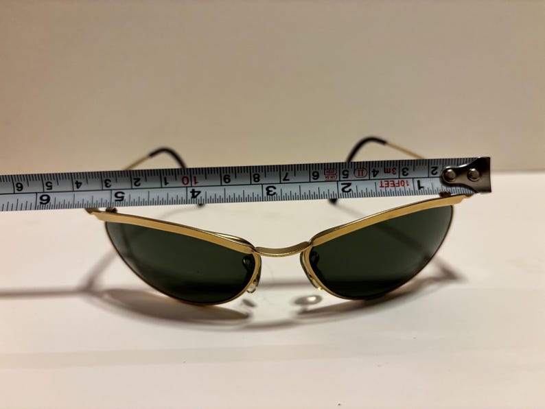 Puede incluir: Gafas de sol con montura dorada y lentes verde oscuro. Las gafas de sol se miden con una cinta m&eacute;trica, mostrando un ancho de aproximadamente 12,7 cm. Las patillas son negras y la montura es dorada.