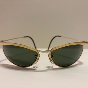 Puede incluir: Gafas de sol con montura dorada y lentes verde oscuro. Las gafas tienen un dise&ntilde;o elegante y curvado con una fina montura dorada y patillas negras. Las lentes son de color verde oscuro, lo que contrasta con la montura dorada.