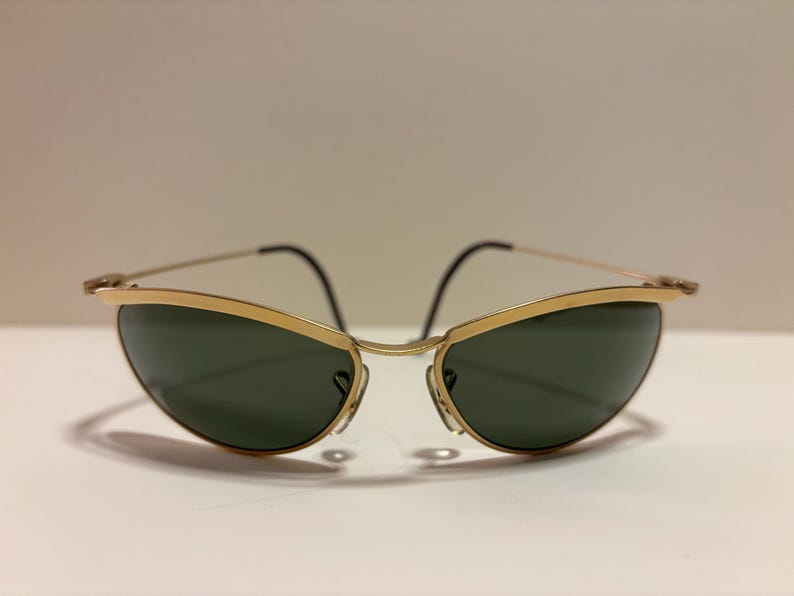 Puede incluir: Gafas de sol con montura dorada y lentes verde oscuro. Las gafas tienen un dise&ntilde;o elegante y curvado con una fina montura dorada y patillas negras. Las lentes son de color verde oscuro, lo que contrasta con la montura dorada.