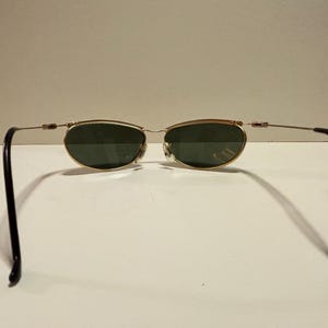 Puede incluir: Gafas de sol ovaladas con montura dorada y lentes verdes. Las patillas son de color marr&oacute;n oscuro. Las gafas est&aacute;n sobre un fondo blanco liso. Las monturas tienen un dise&ntilde;o fino y delicado.