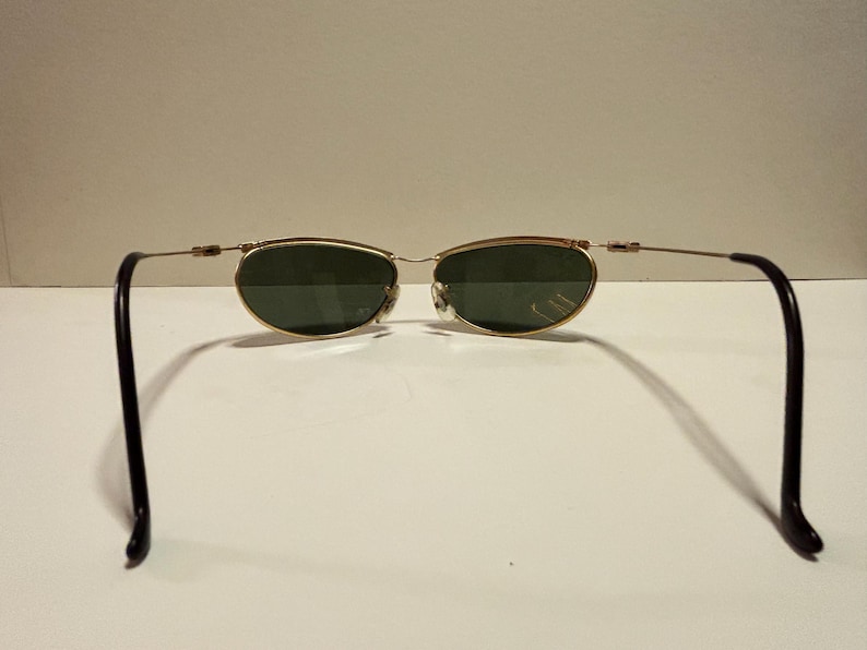 Puede incluir: Gafas de sol ovaladas con montura dorada y lentes verdes. Las patillas son de color marr&oacute;n oscuro. Las gafas est&aacute;n sobre un fondo blanco liso. Las monturas tienen un dise&ntilde;o fino y delicado.