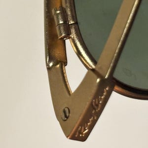 Puede incluir: Primer plano de gafas de sol doradas con lente verde. La montura presenta el logotipo de Ray-Ban. La bisagra y la patilla son visibles, mostrando el dise&ntilde;o y la construcci&oacute;n de las gafas. Las gafas de sol est&aacute;n sobre un fondo blanco.