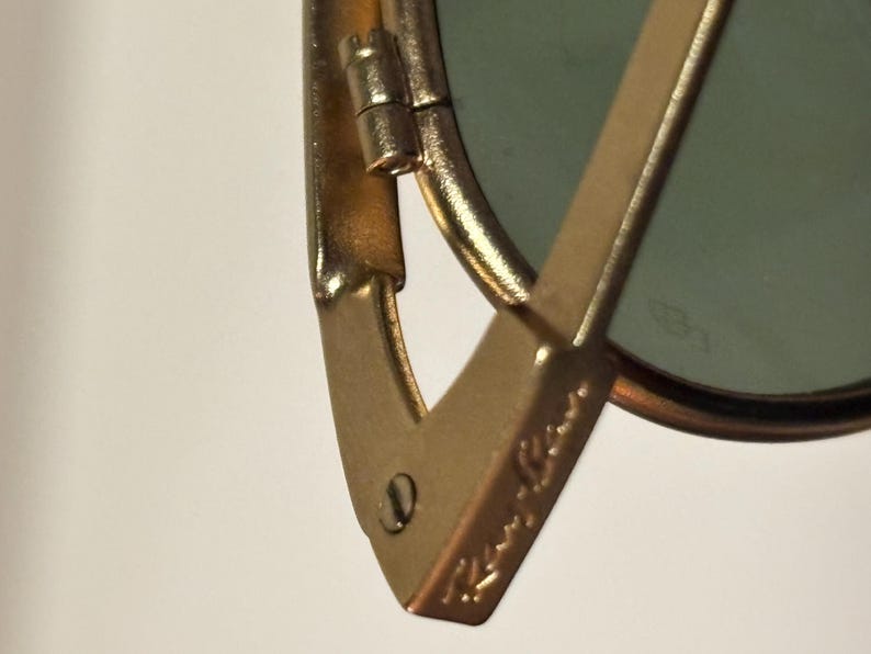 Puede incluir: Primer plano de gafas de sol doradas con lente verde. La montura presenta el logotipo de Ray-Ban. La bisagra y la patilla son visibles, mostrando el dise&ntilde;o y la construcci&oacute;n de las gafas. Las gafas de sol est&aacute;n sobre un fondo blanco.