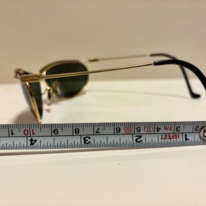Puede incluir: Gafas de sol con montura dorada y lentes verdes, miden aproximadamente 10 cm de largo. Las gafas tienen patillas negras y est&aacute;n sobre una superficie blanca con una cinta m&eacute;trica.