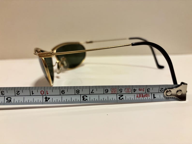 Puede incluir: Gafas de sol con montura dorada y lentes verdes, miden aproximadamente 10 cm de largo. Las gafas tienen patillas negras y est&aacute;n sobre una superficie blanca con una cinta m&eacute;trica.