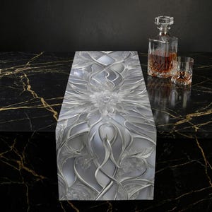 Può includere: Runner da tavolo decorativo con design floreale argento e bianco. Il runner presenta un motivo in rilievo di fiori e viticci. Sullo sfondo sono visibili una caraffa di vetro e un bicchiere da whisky, che suggeriscono un'ambientazione da bar o sala da pranzo.