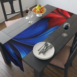 May include: A dark gray wooden dining table is set with a vibrant table runner featuring a blue and red abstract design. The table is set with white plates, silverware, wine glasses, and a bowl of apples.