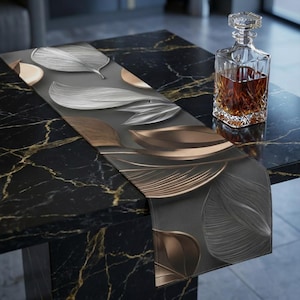 Può includere: Runner da tavolo decorativo con un design a foglie in argento, oro e bronzo su sfondo grigio scuro. Una caraffa di cristallo piena di liquido ambrato è appoggiata su un tavolo di marmo nero.