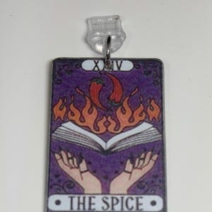 May include: A rectangular pendant featuring a tarot card design. The card has a purple background with an open book, hands, flames, and chili peppers. The text "THE SPICE" is at the bottom. A clear plastic clasp and metal ring are attached at the top.