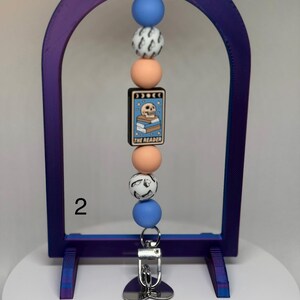 May include: A beaded keychain with a purple arch-shaped frame. The keychain features blue, peach, and patterned beads, and a rectangular charm with a skull on books and the text "THE READER". A silver-colored clip and charm are attached.