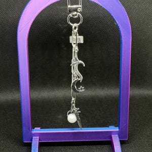 Puede incluir: Un llavero plateado con un dije de espada, luna y estrella, suspendido dentro de un soporte de exhibición en forma de arco púrpura y azul. El soporte tiene un efecto de color degradado y una base rectangular.