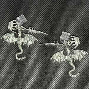 May include: A pair of silver-toned dragon earrings. Each earring features a detailed dragon with outstretched wings, a sword, and a crown-like top. The earrings have a post-back closure and are displayed against a dark background.
