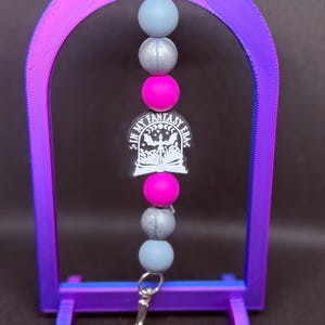 Puede incluir: Un cordón de cuentas con un cierre plateado y un dije que dice "IN MY FANTASY ERA". El cordón presenta cuentas grises, plateadas y rosa intenso. El soporte en forma de arco es morado y azul.