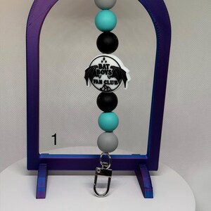 May include: A beaded lanyard with a silver clasp and a "Bat Boys Fan Club" emblem. The lanyard features gray, teal, and black beads. The emblem is white with black bat wings and text. The number "1" is visible in the image.