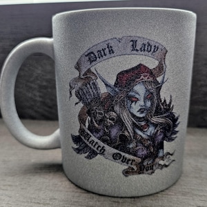 May include: A silver ceramic mug with a white handle. The mug features a graphic of a female fantasy character with red eyes, wearing a red hat, and the words "Dark Lady" and "Watch Over You" on a banner.