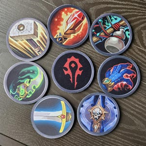 Puede incluir: Nueve posavasos redondos con varios diseños de fantasía. Los posavasos presentan imágenes de un martillo, una espada, una mano con llamas y otros símbolos. Cada posavasos tiene un borde gris oscuro y está sobre una superficie de madera.