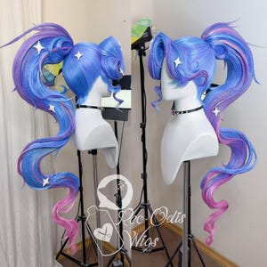 Op de afbeelding: Een blauw, paars en roze pruik met staartjes en sterversieringen. De pruik wordt getoond op een mannequinhoofd met een zwarte choker. Het haar is in lange staarten gevlochten, ideaal voor cosplay of thema-evenementen.
