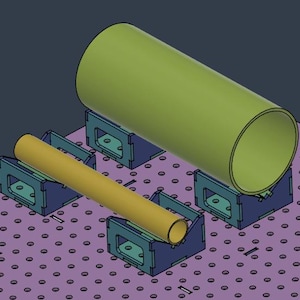 Może przedstawiać: Cyfrowe renderowanie konfiguracji warsztatowej z dużym, oliwkowozielonym obiektem cylindrycznym i mniejszym, żółtym obiektem cylindrycznym. Oba są podparte niebieskimi i turkusowymi wspornikami na fioletowej perforowanej powierzchni.