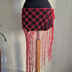 Puede incluir: Una falda de macramé roja con un patrón geométrico y flecos largos se exhibe en un maniquí negro. La falda está hecha de cordón rojo anudado, creando un diseño decorativo de tejido abierto. La falda se exhibe en un soporte de madera.