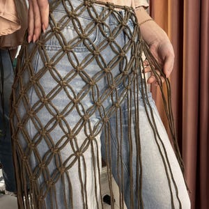 May include: A brown macrame belt featuring a diamond-shaped knotted grid pattern and long, dangling fringe strands. The belt is worn over light blue denim jeans, showcasing the intricate cord construction and the open-weave design.