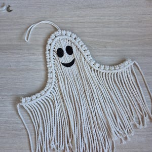 May include: A macrame ghost wall hanging crafted from off-white cotton cord. The ghost features a rounded head with two black felt oval eyes and a black felt curved mouth. The lower portion consists of long, loose hanging fringe strands.