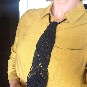 May include: A black macrame necktie featuring an intricate knotted geometric pattern. The tie is constructed from cord and hangs from a thin cord loop that secures around the neck, displaying a textured, woven design throughout the length.