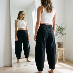 Op de afbeelding: Een persoon draagt een witte tanktop en een zwarte spijkerbroek met een elastische tailleband. De broek heeft een unieke, volumineuze vorm. De foto is genomen voor een grote spiegel, die de outfit weerspiegelt.