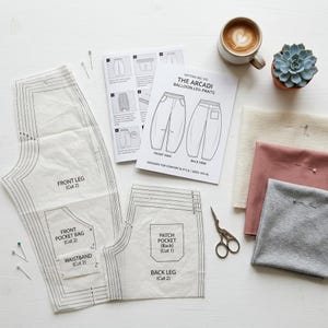 Op de afbeelding: Een bovenaanzicht van naaibenodigdheden, waaronder patroondelen, stofstalen en een schaar. Het patroon is voor "THE ARCADI BALLOON-LEG PANTS". Een kopje koffie en een kleine vetplant zijn ook aanwezig.