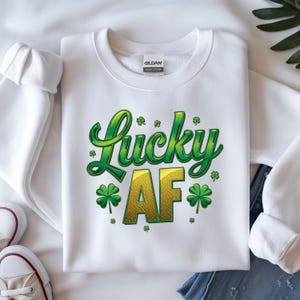 Könnte beinhalten: Weißes Sweatshirt mit dem Schriftzug "Lucky AF" in Grün und Gold, verziert mit Kleeblättern. Das Sweatshirt besteht aus weichem Material, ideal für St. Patrick's Day.
