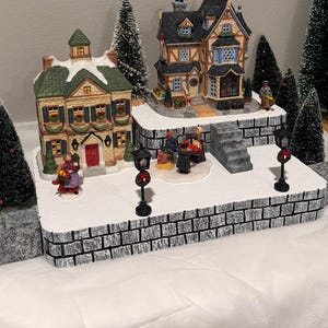 May include: A Christmas village scene featuring two detailed houses, miniature evergreen trees, and figures. The houses have snow-covered roofs and are decorated with festive garlands. The scene is set on a platform with a brick-like facade.