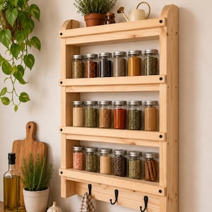 May include: A light-coloured wooden wall-mounted spice rack featuring three horizontal shelves filled with glass jars containing various colourful spices, and three black metal hooks attached to the bottom frame for hanging kitchen items.