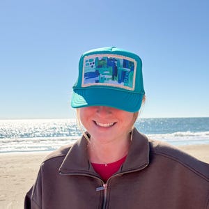 May include: A teal trucker hat with a rectangular patch featuring an abstract design in shades of blue and green. The hat has a mesh back and a curved brim. The person is wearing a brown zip-up jacket and a red shirt.