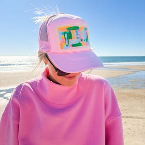以下が含まれることがあります： メッシュバックとカラフルな抽象的なデザインパッチが付いたピンク色のトラッカーハット。 ピンクのスウェットシャツとサングラスを着用した人が帽子をかぶっており、背景にはビーチと海があります。