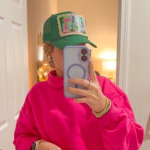 May include: A green trucker cap with a rectangular patch displaying the letters "TA" in pink and green. The person is wearing a vibrant pink sweatshirt and holding a light blue phone.