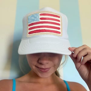 Puede incluir: Gorra de camionero blanca con un diseño de bandera americana pintada a mano. La bandera tiene rayas rojas y un cuadrado azul con estrellas blancas. La gorra tiene una visera blanca y una parte trasera de malla.