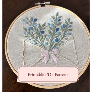 May include: Embroidered hoop art featuring a floral design. The artwork showcases a light-colored envelope with a bouquet of blue and green embroidered flowers. A pink bow adorns the envelope. The text "Printable PDF Pattern" is displayed on a pink banner.