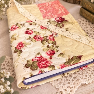 Peut inclure: Une couverture de livre en tissu matelassé présentant un motif patchwork de tissu crème et des imprimés floraux avec des roses rouges. La couverture comprend une bordure en dentelle crème et est cousue avec un motif matelassé ondulé.