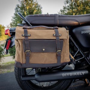 May include: A brown and tan motorcycle saddlebag with leather straps and buckles is attached to a black motorcycle. The bag has a flap closure and a handle. The motorcycle has a black exhaust pipe with the text "DIYBIKELY".