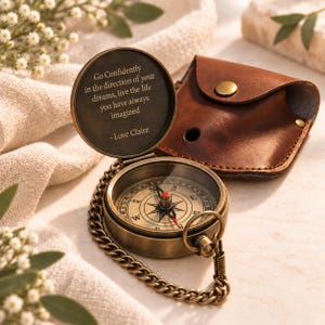 Puede incluir: Una brújula de bolsillo de latón con cadena y una funda de cuero marrón. La tapa está abierta, revelando el texto grabado: Go Confidently in the direction of your dreams, live the life you have always imagined - Love Claire. La esfera muestra los puntos cardinales.