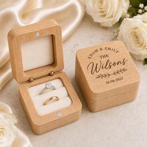 Könnte beinhalten: Eine quadratische Ringbox aus hellem Holz mit Klappdeckel, graviert mit dem Text "ADAM & EMILY THE Wilsons 16.06.2027" und einem Blattmotiv. Das offene Kästchen zeigt ein weißes Samtfutter mit einem silbernen Diamantring und einem Goldring.