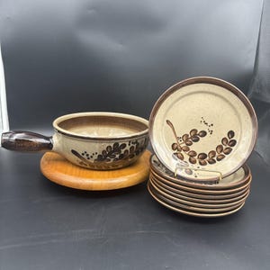 May include: A ceramic soup bowl with a handle and a stack of matching plates, all featuring a speckled beige glaze with a dark brown painted leaf pattern and a solid brown rim. The bowl rests on a circular wooden base, while the plates are stacked.