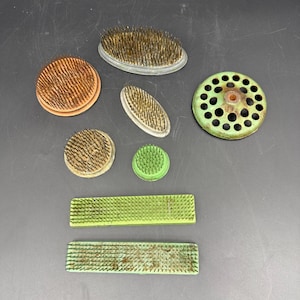 May include: A collection of vintage flower frogs in various shapes and sizes. The frogs include round, oval, and rectangular designs, with metal pins for arranging flowers. Colors include green, orange, and silver.